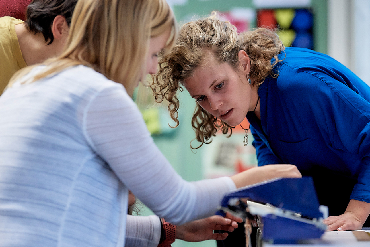 Ein Seminar im Textilen Gestalten. Eine Dozentin zeigt, wie eine Strickmaschine funktioniert, zwei Studierende schauen zu.