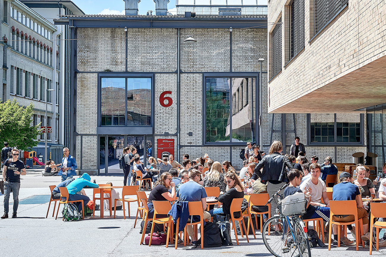 Studierende und weitere Personen sitzen an Tischen vor dem Hochschulgebäude vonRoll.