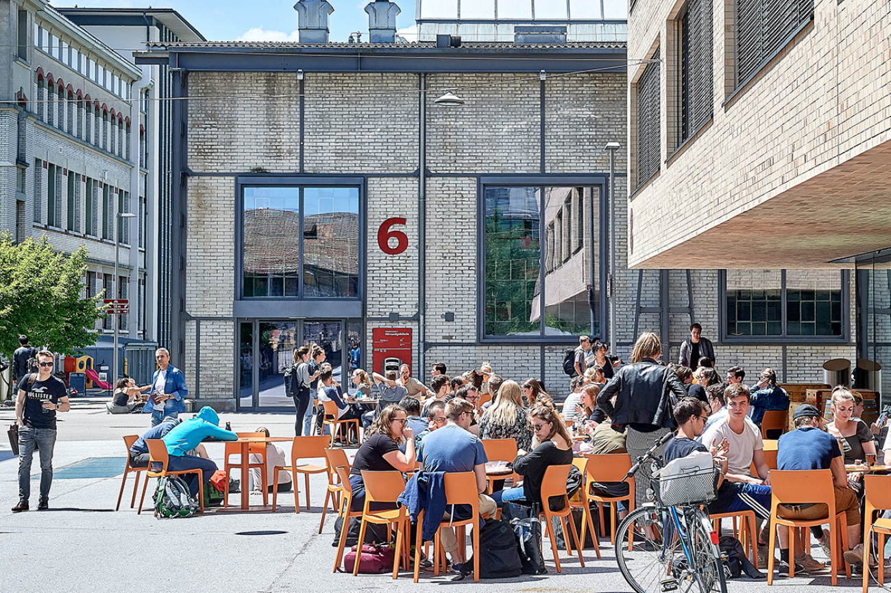 Ein Bild des vonRoll-Campus. Studierende sitzen an runden Tischen vor dem Gebäude an der Fabrikstrasse 8.