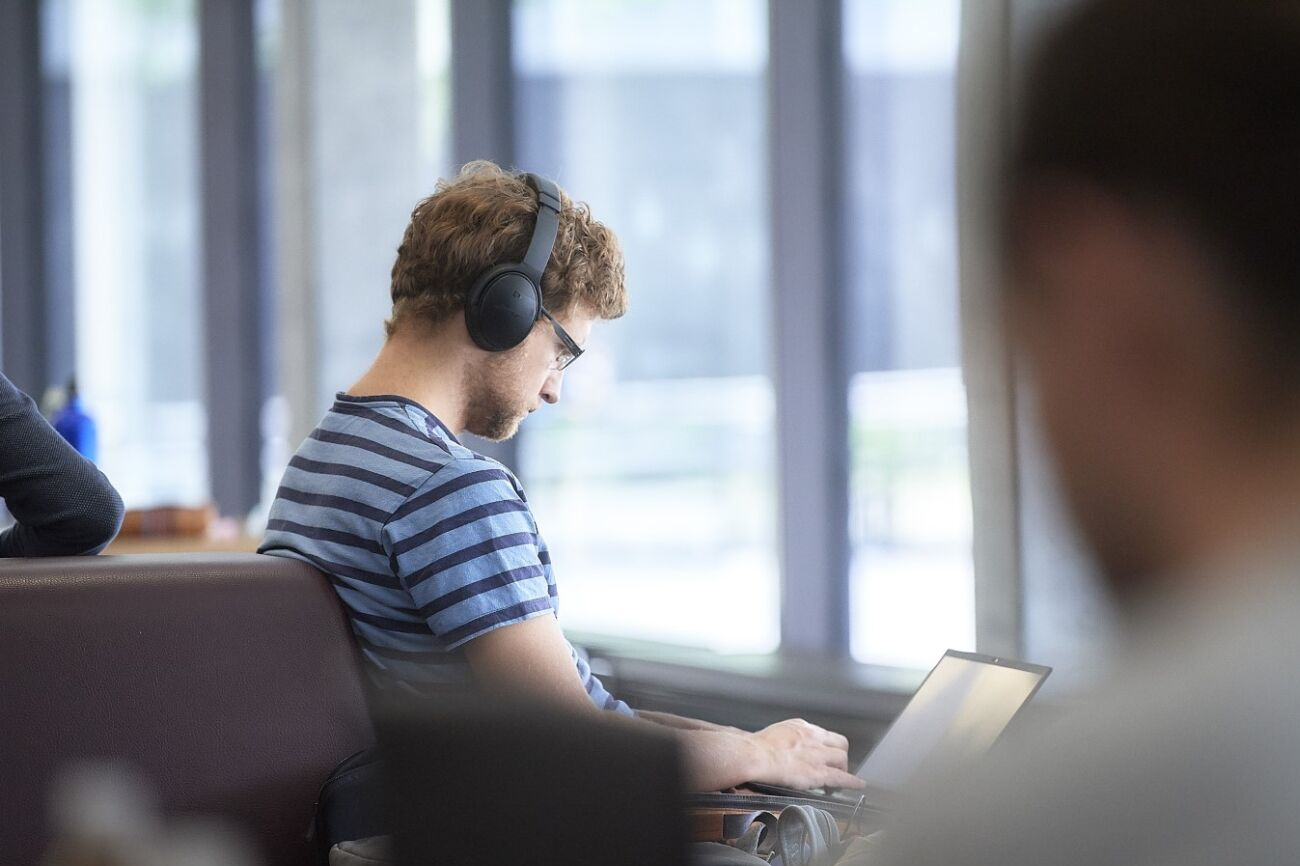 Ein junger Mann in T-Shirt und einem Kopfhörer auf dem Kopf arbeitet auf seinem Laptop-Computer in der Lounge des PHBern-Gebäudes