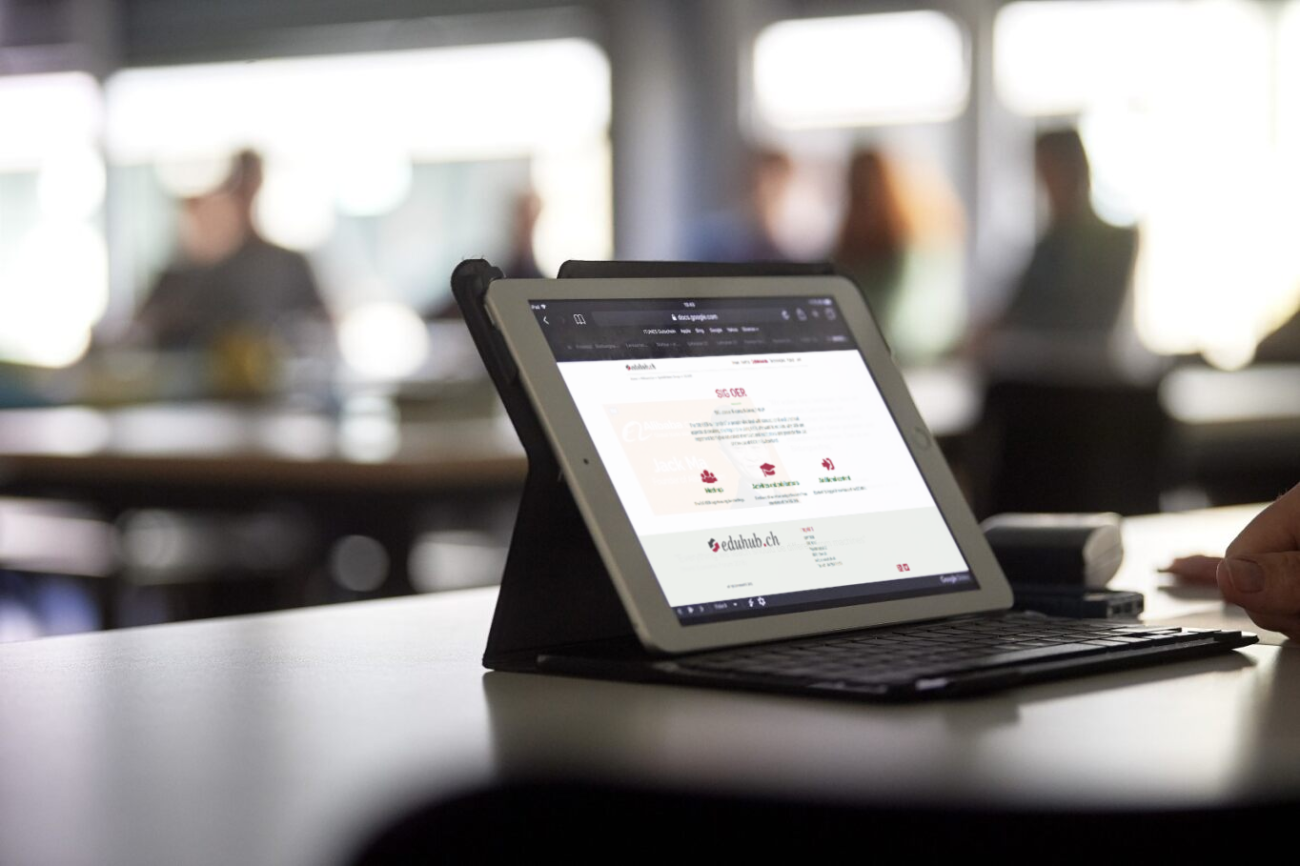 Ein Sitzungszimmer. Im Vordergrund scharf ein Tabletcomputer. Im Hintergrund sind sehr unscharf Personen zu sehen, die arbeiten.