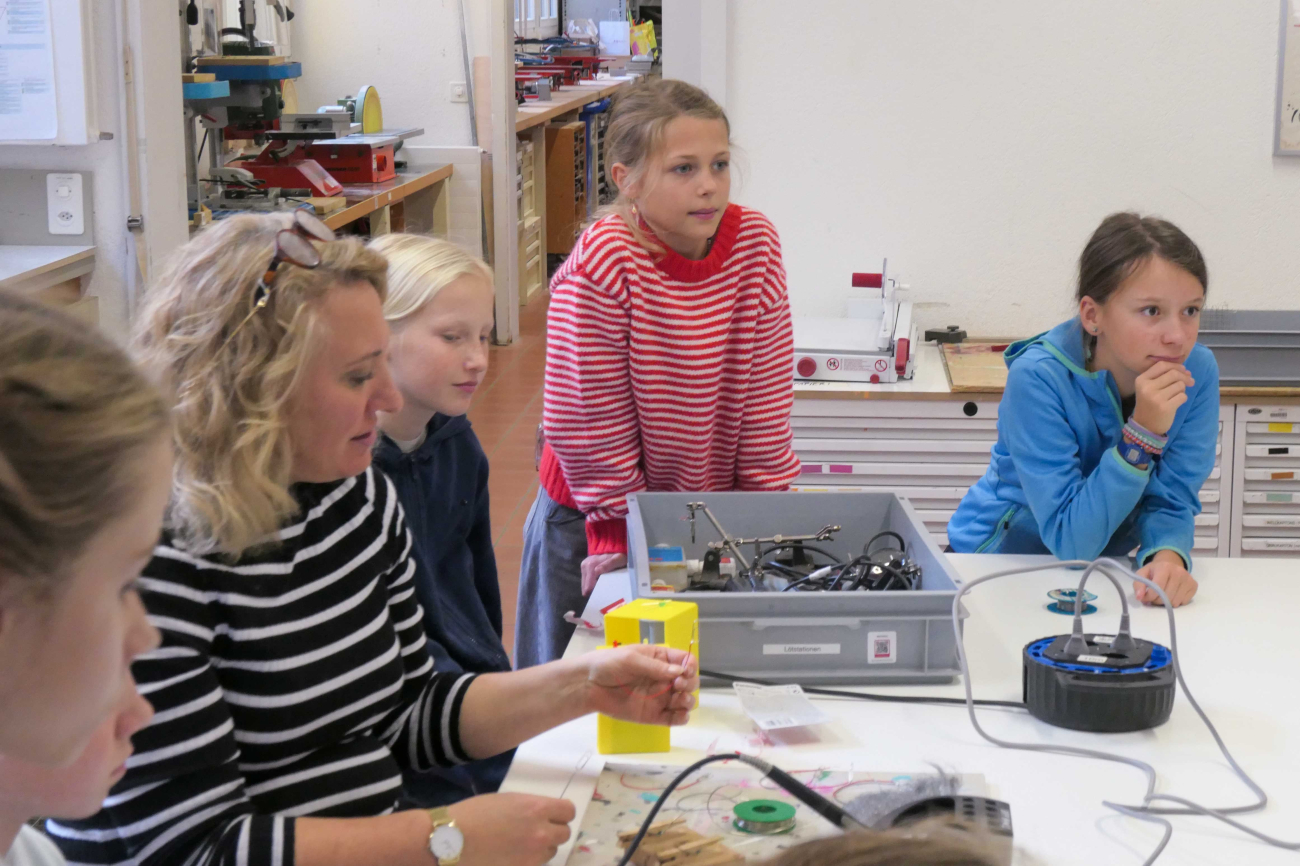 Mädchen schauen gespannt in eine Richtung, eine Frau macht etwas auf einem Tisch