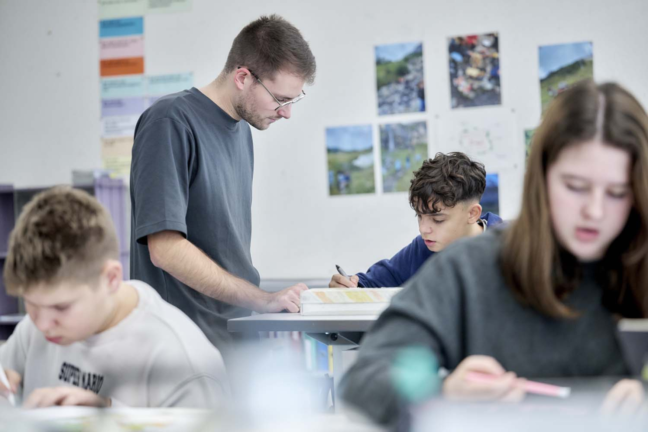 Junger Lehrer schaut an Pult eines Schülers auf dessen Arbeitsblatt
