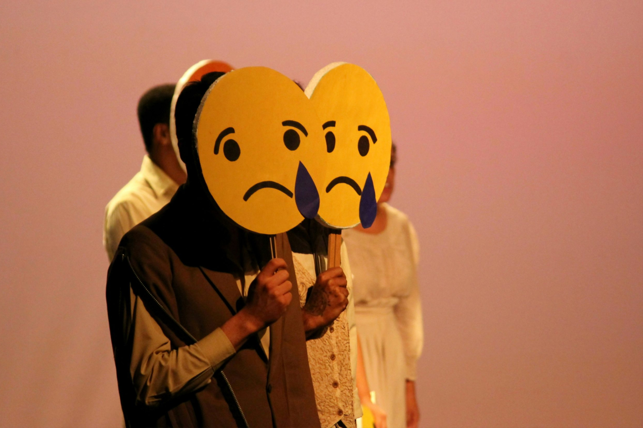 Theaterdarsteller:innen halten trauriges Smiley-Gesicht aus Karton vor das Gesicht