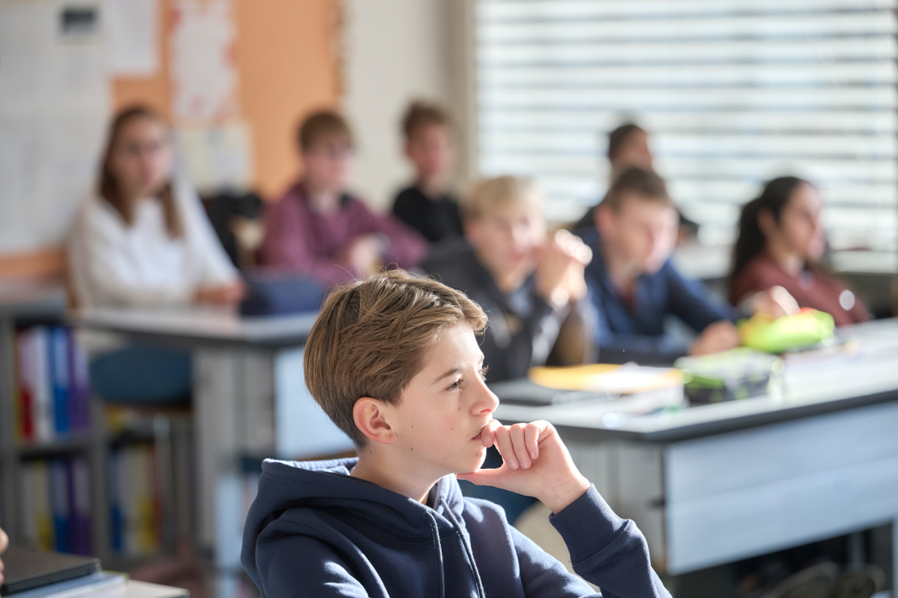 Jugendlicher nachdenkend im Unterricht, in der Hintergrundunschärfe viele seiner Klassenkameraden