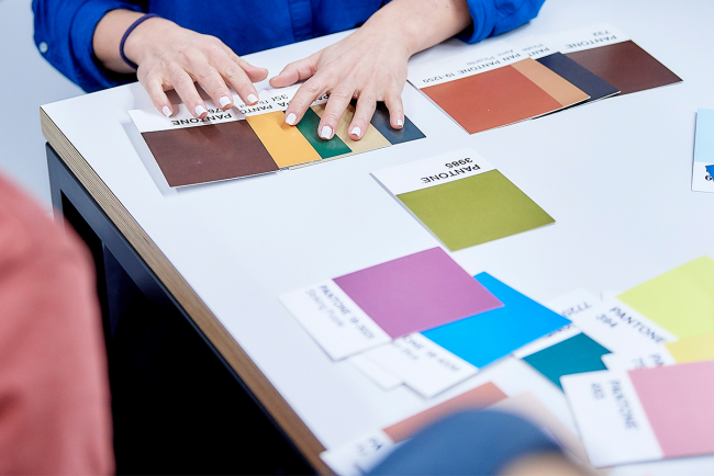 Mehrere Personen sitzen um einen Tisch, auf dem Pantone-Farbkarten aufgelegt sind. Eine Studentin schiebt verschiedene Farben zusammen.