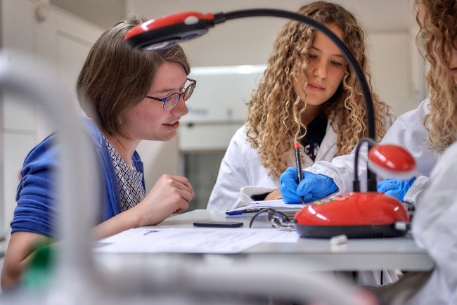 Eine Praktikantin unterrichtet Biologie an einer Fachmittelschule im Labor. Sie betrachtet ein Blatt, auf dem eine Schülerin etwas schreibt. Im Hintergrund sind weitere Schülerinnen zu sehen, die weisse Kittel tragen.