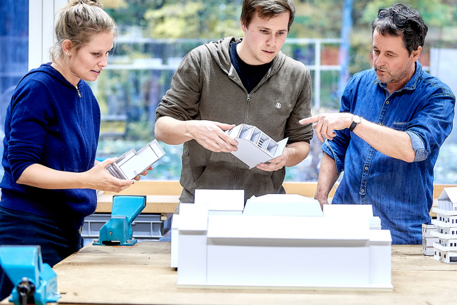 Eine Studentin, ein Student und ein Dozent betrachten zusammen ein Architekturmodell.