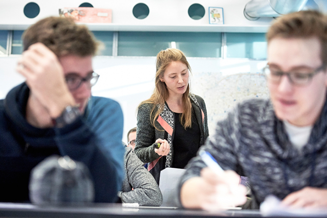 Eine Studentin des Instituts Sekundarstufe II im Praktikum. Sie steht im Klassenzimmer zwischen Schülern und hält einen Leuchtstift in der Hand. Im Vordergrund sind unscharf zwei Schüler zu erkennen.