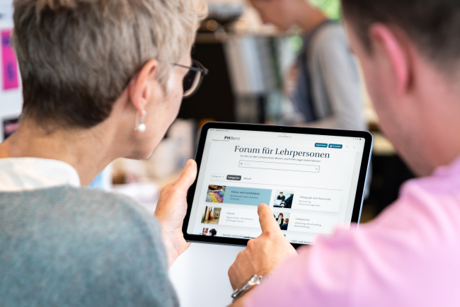 Frau und Mann schauen gemeinsam auf ein Tablet, zu sehen ist der Screen des Forums für Lehrpersonen