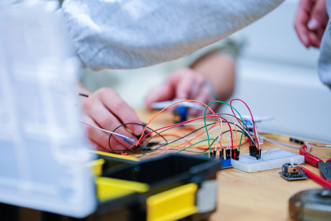 Ein Tisch, auf dem ein Zange, ein Werkzeugkasten und eine Microcontroller liegt, zu dem viele bunte Drähte führen.