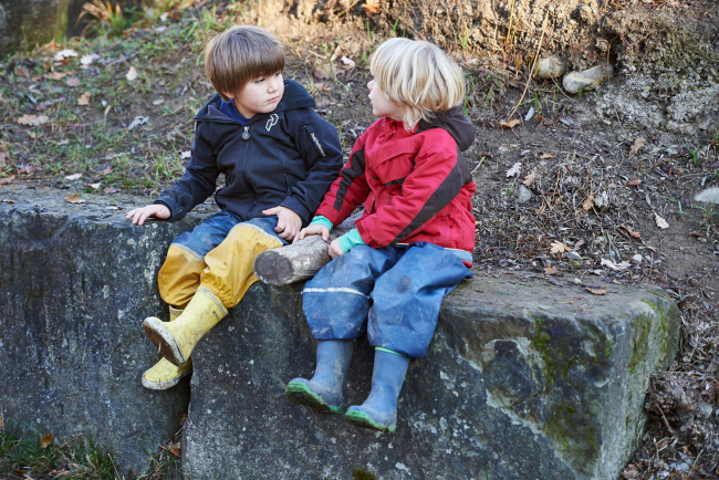 zwei Kinder, die mit Gummistiefeln auf einer Mauer sitzen