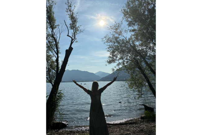 Lara streckt die Hände aus. Im Hintergrund ist ein See, Links und rechts von Lara sind Bäume.